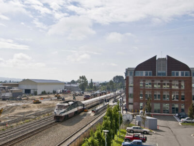 A drone view of the train passing through Everett Station. The 4-story building is on the left and in the background is a barn with farm implements around it.