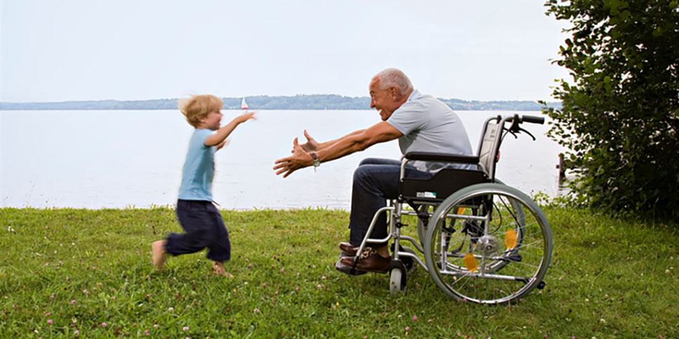 A child runs through the grass to greet an older person who is sitting in a wheelchair. The older person is leaning forward with their arms spread ready to give the child a hug.