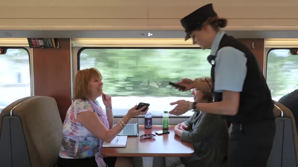 Two people sit across from each other in a train car. A train employee is using a handheld device to scan the passengers' ticket information from their phones.