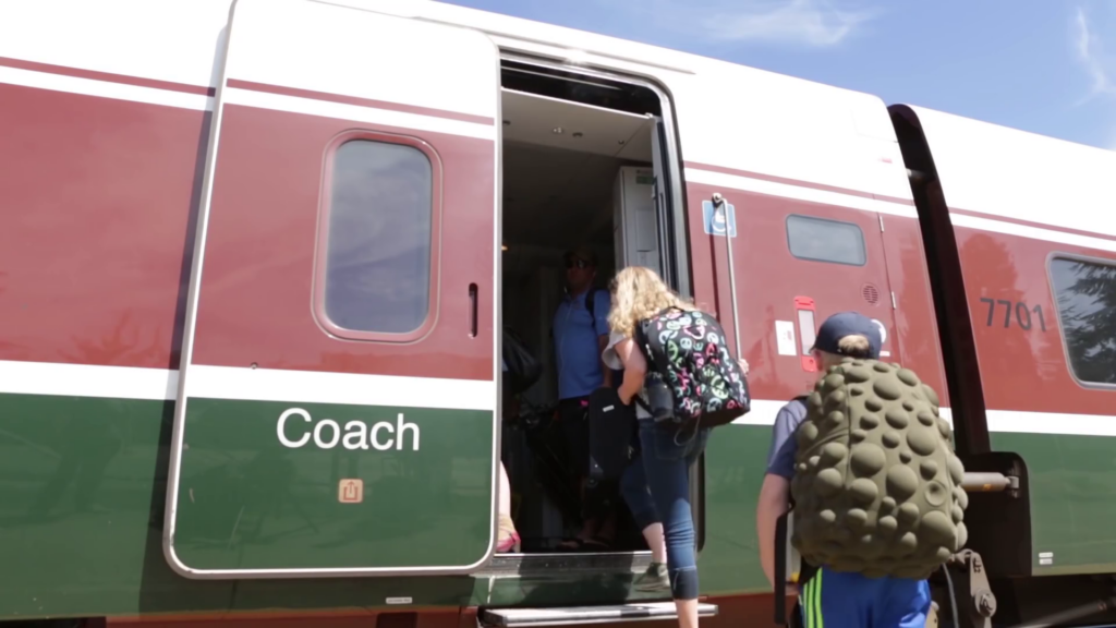 Two people carrying decorative backpacks step aboard a train. The open door to the train has the word Coach painted on it.