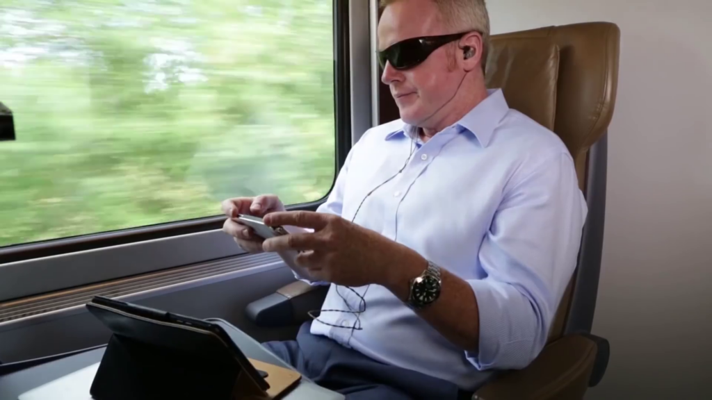 A person sits next to a window inside a moving train. They are wearing sunglasses, listening to headphones, and are looking at their mobile device.