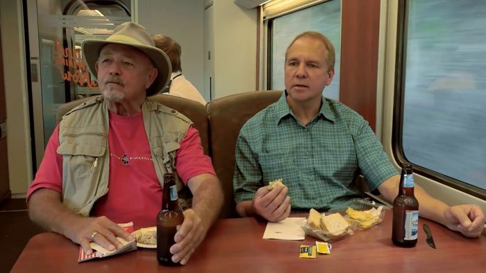 Two people sit at a table in a train car. They each have a beverage in a glass bottle and a lunch in front of them.