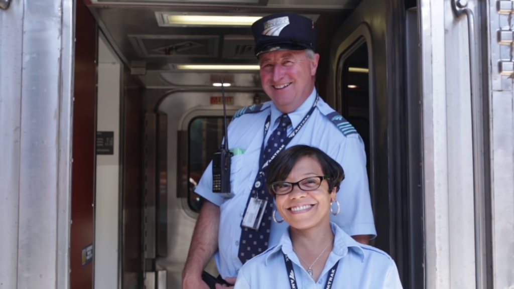Two train workers stand in the open door of a train car, smiling at the camera.
