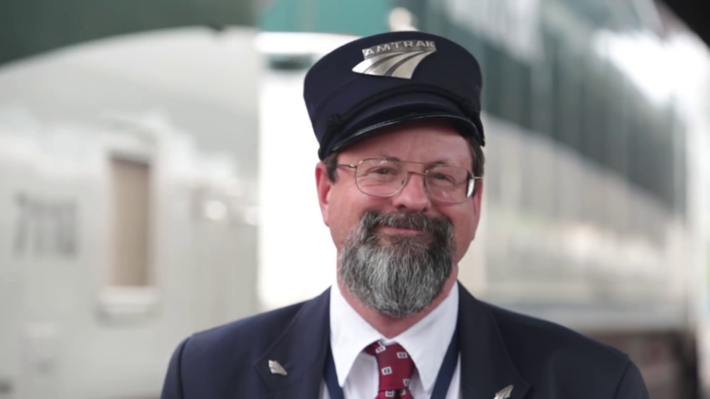 A friendly-looking man wearing a hat with the Amtrak logo on it smiles at the camera.