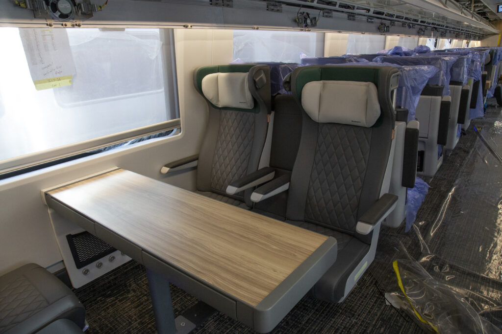 Two seats in the interior of the train. The head rest has text printed on it that says "Business." There is a table in front of the two seats. The rest of the car is visible in the background with protective plastic still on the rest of the seats.
