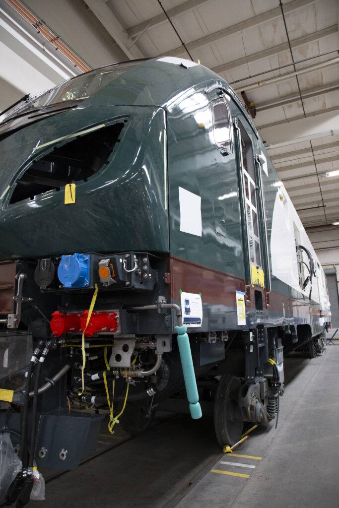 The front of a train car in a warehouse. The car is a dark green with white accents.