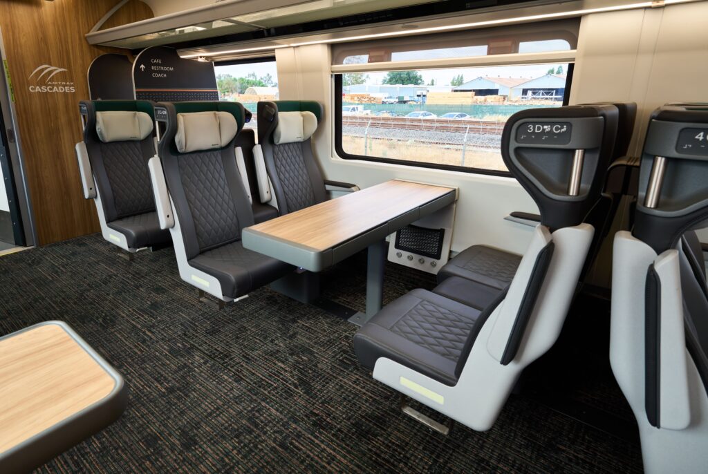 Interior photo of a new Amtrak Cascades Business Car showing two seats on one side of the car. The leather seats appear comfy with tall, puffy headrests. Some seats are facing and have tables between them. You can see the edge of a table on the single seating opposite side of the car. The windows are large to view the spectacular Northwest scenery. The floors are carpeted. The end wall has wood paneling with a white Amtrak Cascades logo.