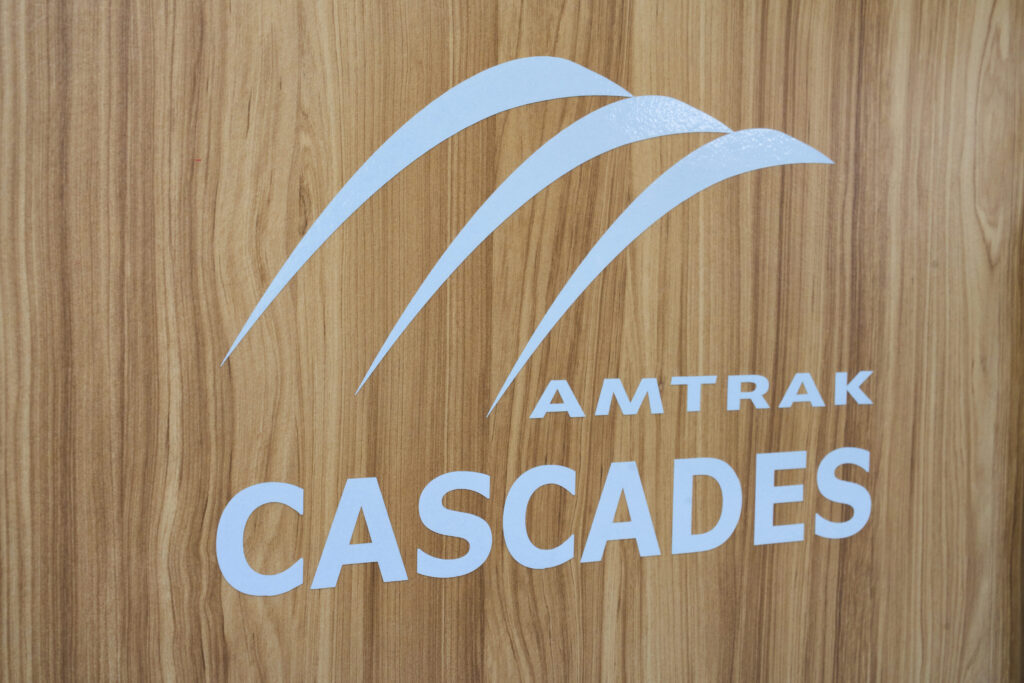 Photo of a white logo with the three mountains above the stacked Amtrak Cascades all caps letters on wood paneling.
