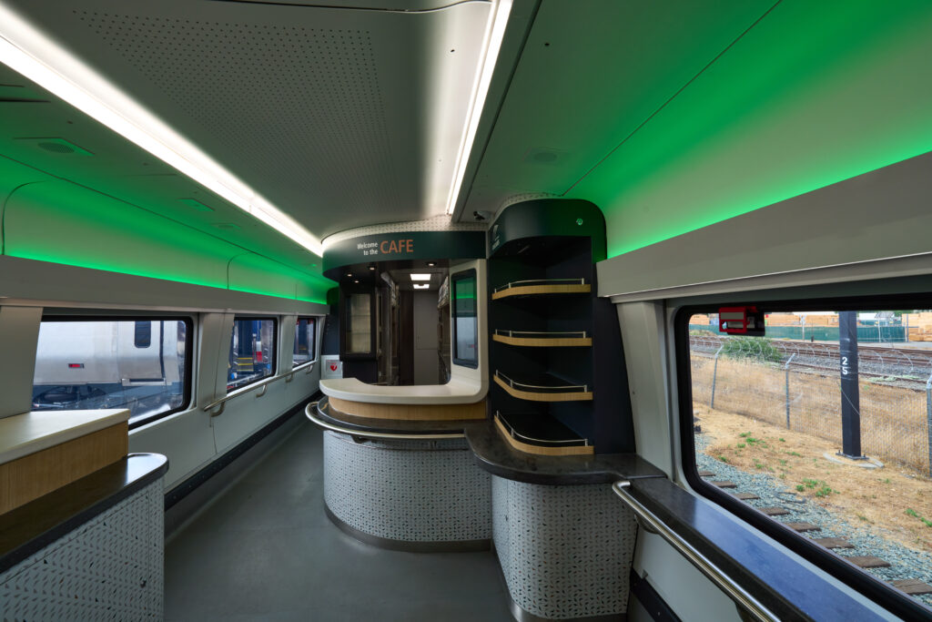 New spacious Amtrak Cascades Cafe Car features windows on both sides. A large service counter sits in the middle. Evergreen and cream colors with gold accents match the Amtrak Cascades logo, a reflection of the Pacific Northwest. White and green shadow lighting create lovely hues on the ceiling.