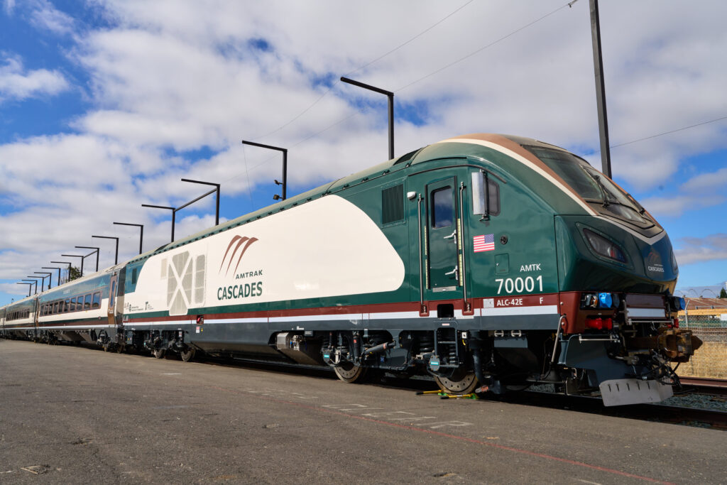 A newly manufactured Amtrak Cascades locomotive waits to leave the factory in California. The frame around the windshield is gold. The nose is evergreen to represent the PNW trees and most of the locomotive is cream, the white color representing the cream in your coffee. The updated Amtrak Cascades logo is large inside the cream portion. Blue sky and thin white clouds create the backdrop.
