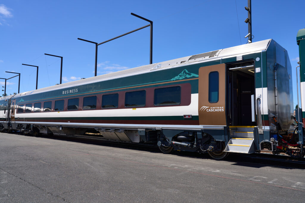 A new Amtrak Cascades Business Train Car waits to leave the manufacturing facility for testing. Cascade mountains are featured on both ends of the car. BUSINESS is printed on the top middle portion of the evergreen background. The mocha color frames the plentiful windows and there's a stripe of cream color below the windows. The Business Car sports a gold door which matches the mountains in the Amtrak Cascades logo. The door is open and the steps are down, waiting for you to hop onboard!