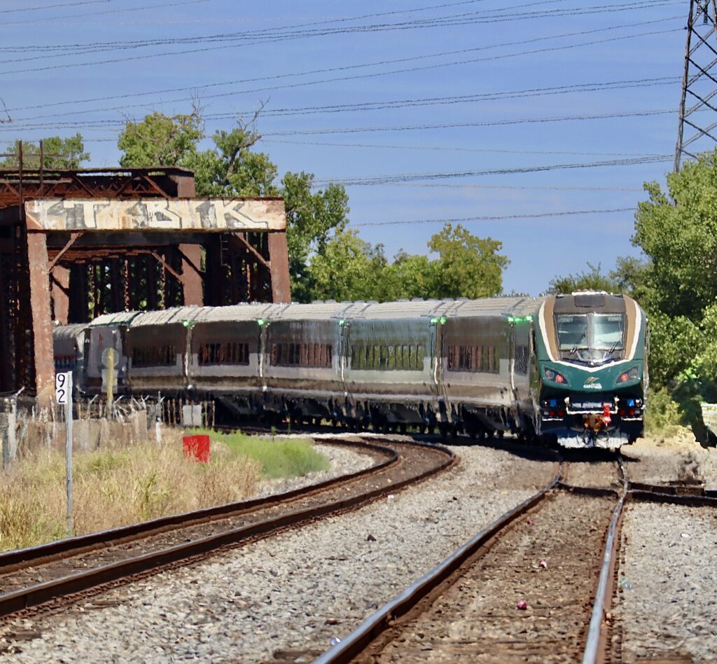 New Amtrak Cascades train near Elvas/Sacramento, CA, rounds a bend under a rail bridge steel structure.