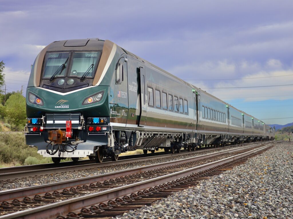 A new Amtrak Cascades train on the tracks with green marker lights at the top ends of each train car as the new train makes it way to testing in Colorado. The cab car is clearly visible on the end of the train while the other cars blur into the remaining light.