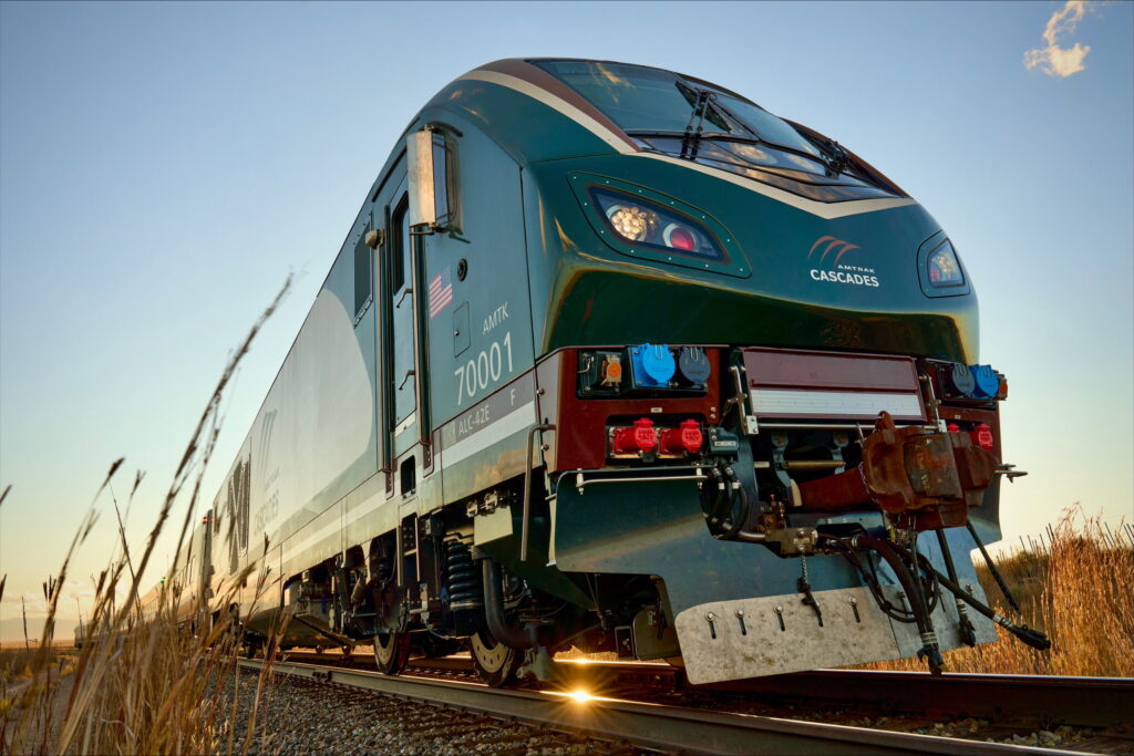 The evergreen, mocha and cream-colored nose of the new Amtrak Cascades Trainset #1 looms large under a dreamy blue sky on the tracks with some tall grasses on either side of the photo. The windshield is accented with the gold nugget color and the Amtrak Cascades logo adorns the evergreen-colored space between the headlights.