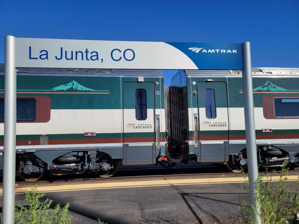 The train station signpost at La Junta, Colorado, frames the new Amtrak Cascades Trainset #1 as it passes through La Junta on its way to Chicago and then Philly. You can see two passenger coach doors and some signature Cascades Mountains accenting the evergreen, mocha, and cream Pacific Northwest-inspired color scheme.