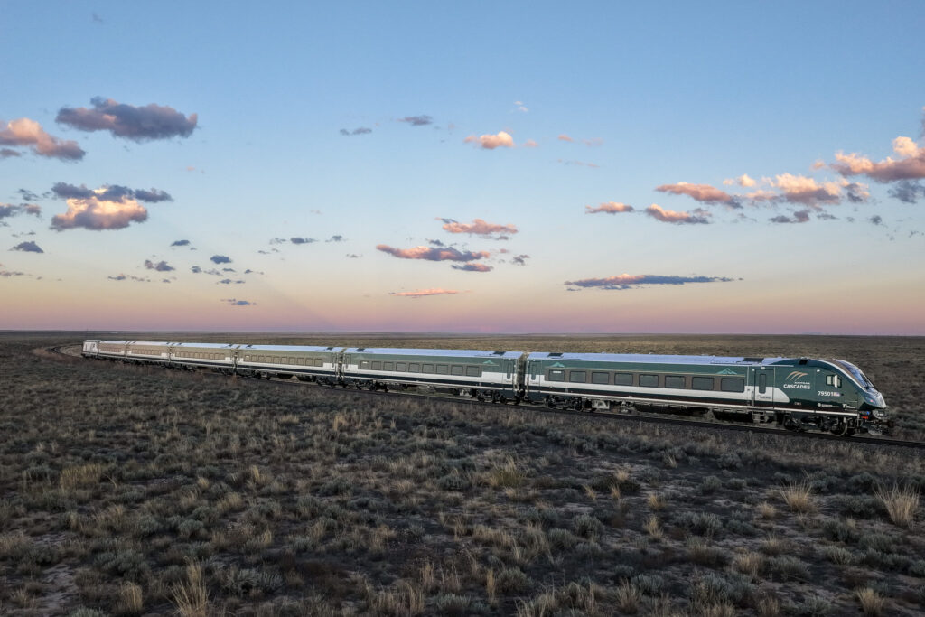 Photo of the new Amtrak Cascades trainset with the cab car in the lead with sagebrush in the foreground and lovely blue sky with some dashes of small clouds above. Photo courtesy of Siemens.